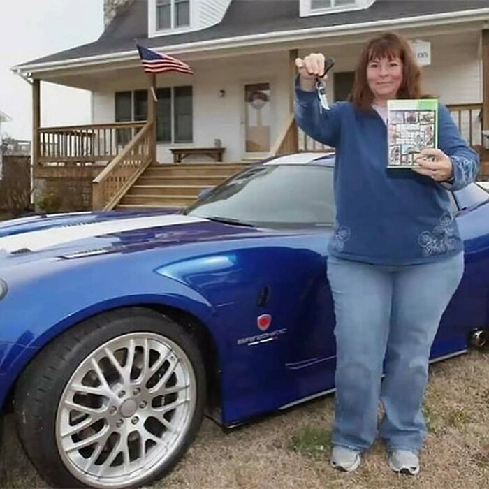 Woman holding car keys and a manual, standing next to a blue sports car in front of a house in a nature setting.