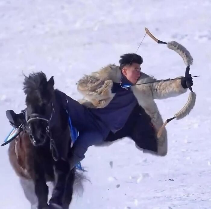 Man wearing fur coat aiming bow and arrow while riding a black horse in snowy nature with animals around.