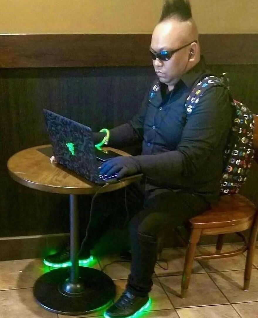 Man with mohawk and sunglasses using laptop, wearing glowing shoes and gloves, showcasing bizarre yet hilarious style.