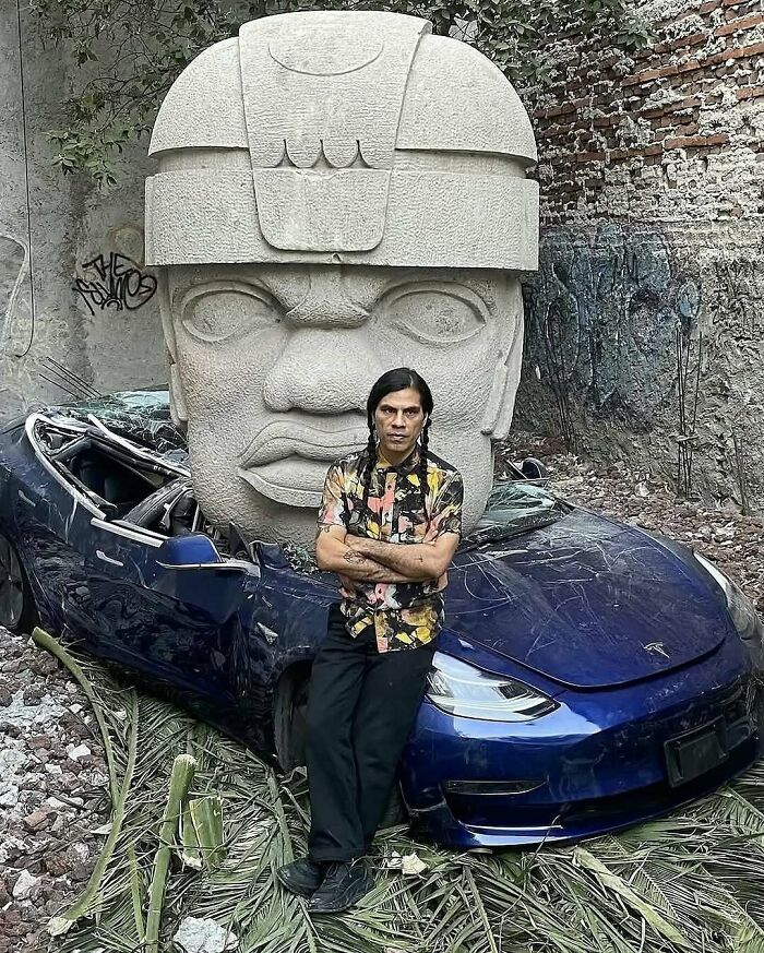 Man leaning against a damaged blue car in front of a large stone sculpture, showcasing bizarre nature and animal facts.