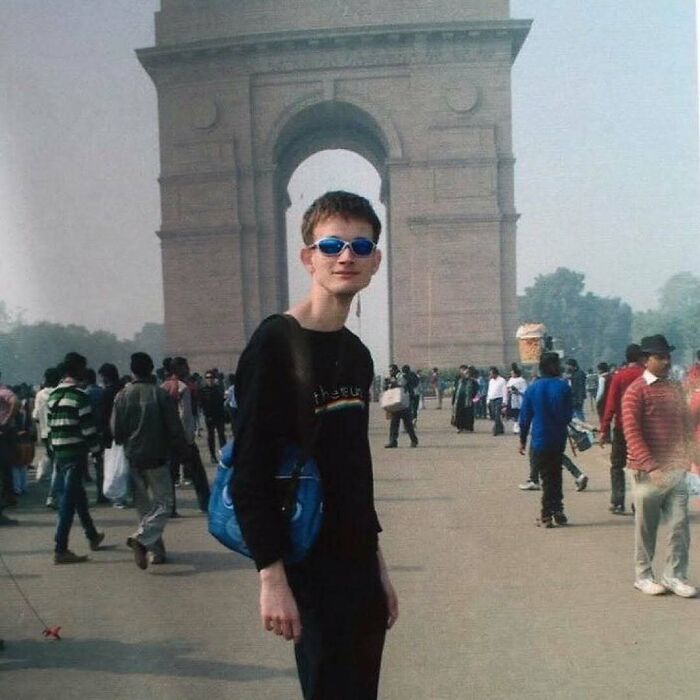 Young man with sunglasses and a blue bag standing in front of a large arch, with a crowd in the background, nature and animals context.