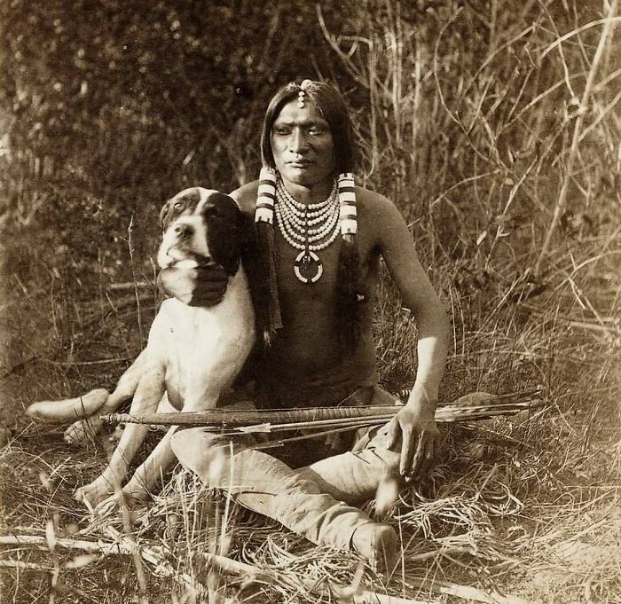 Vintage sepia photo of a Native American man sitting in grass with a dog and arrows, illustrating weird things about history.