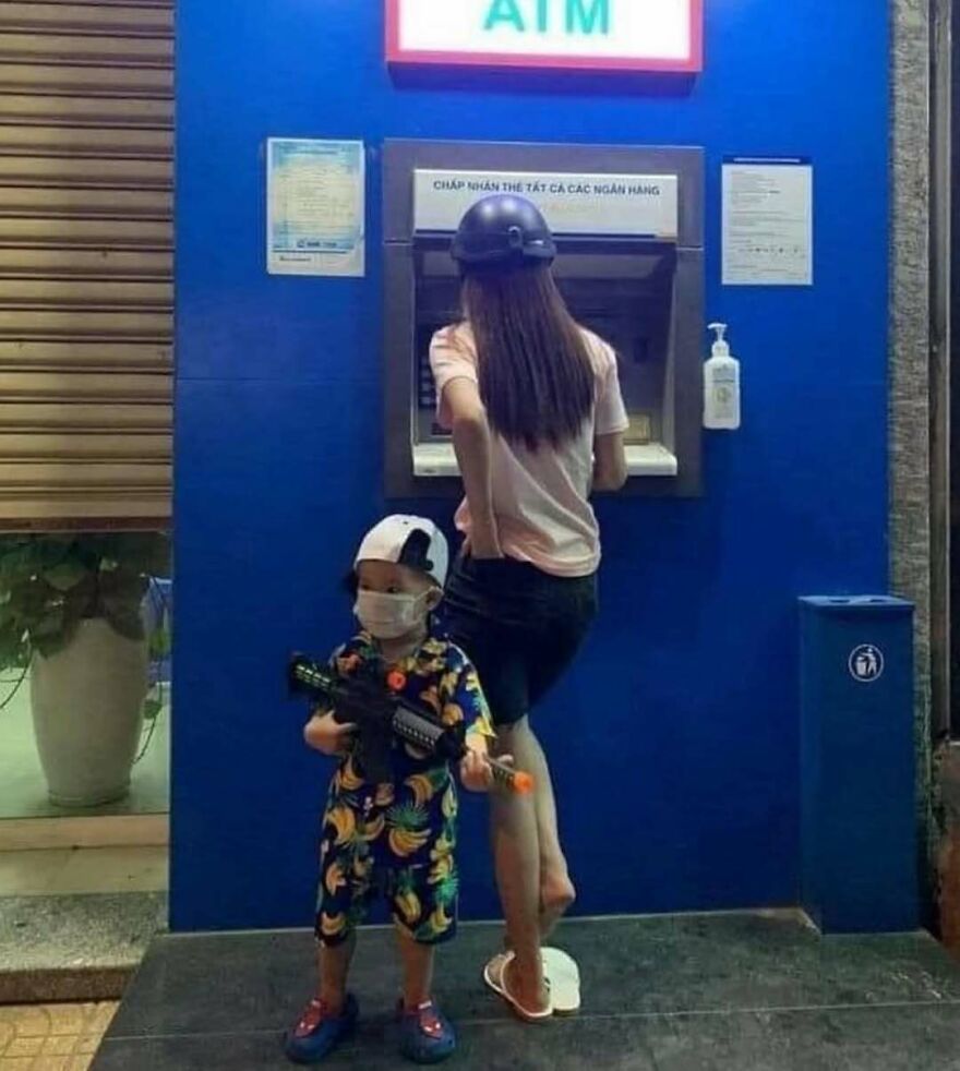 Child wearing a mask and holding a toy gun stands near a woman at an ATM in a bizarre yet humorous scene.