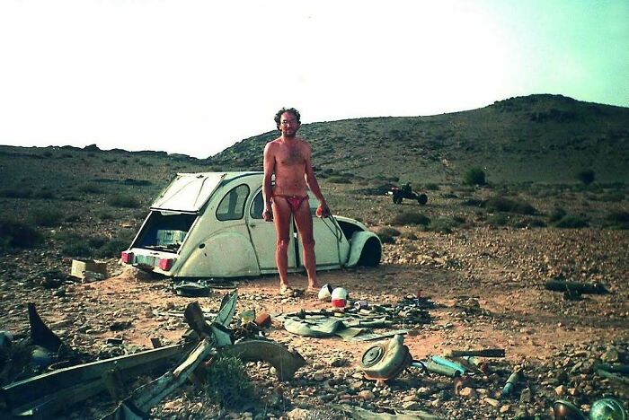 Man in swimwear standing by vintage car wreck in a desert landscape, historical scene from confusing old photos.