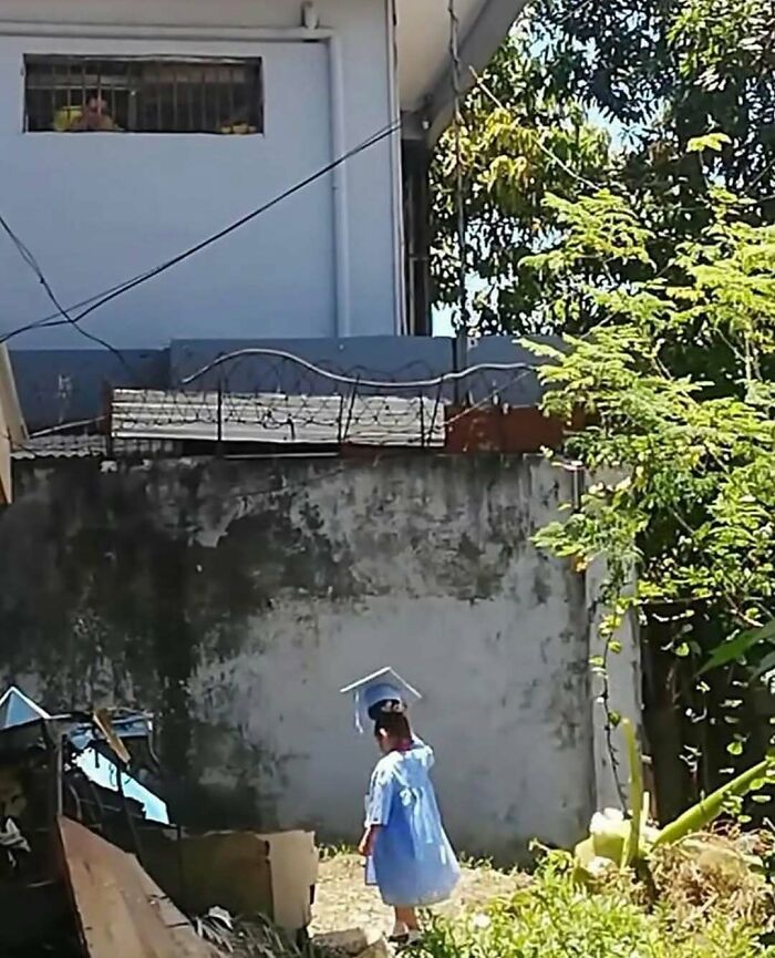 Child wearing a blue graduation gown and cap walking outside near a weathered wall with green trees in nature surroundings.