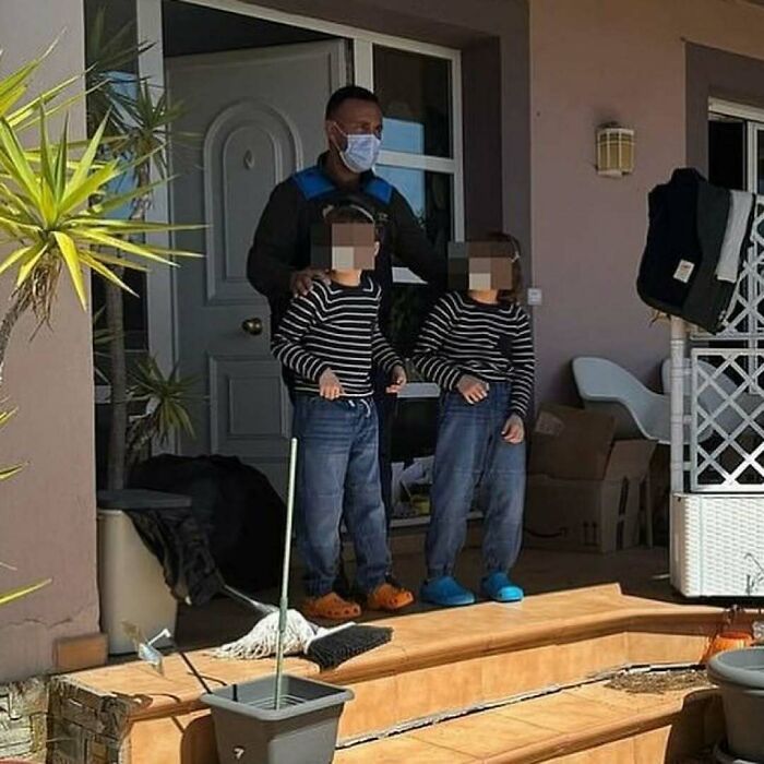 Man wearing mask stands with two children in matching striped shirts and shoes on a porch in a nature setting.