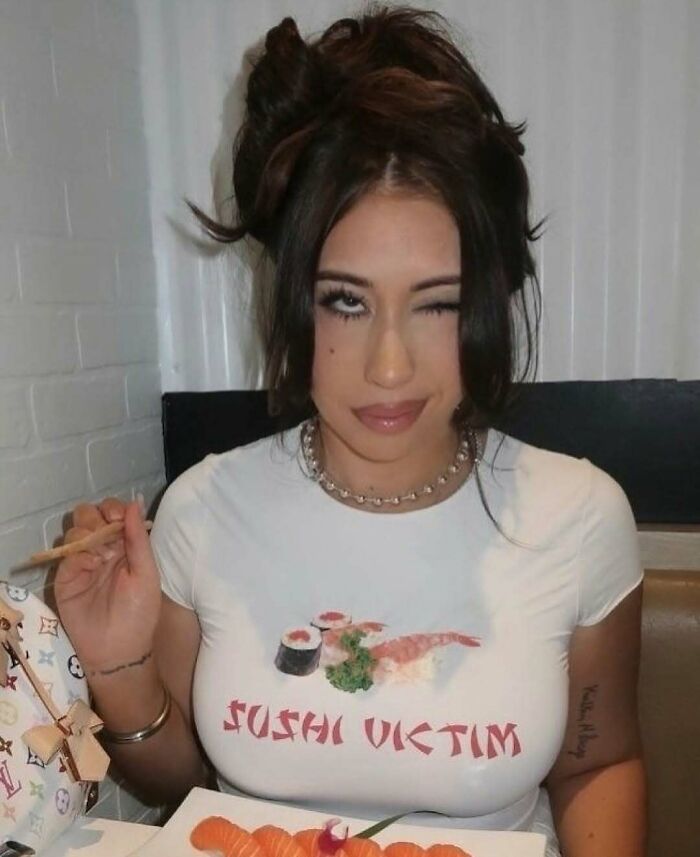 Young woman wearing a sushi victim shirt, holding chopsticks, dining indoors with a plate of sushi in front of her.