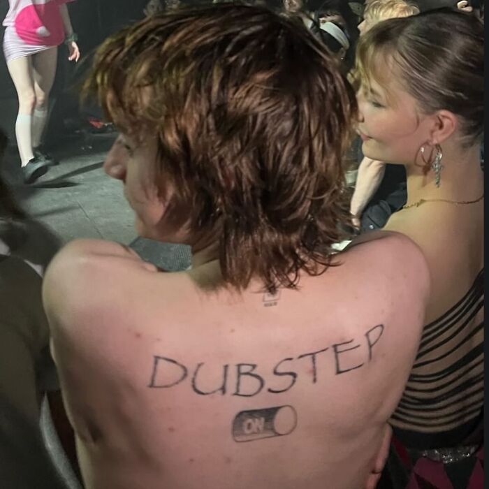 Person with bizarre tattoos on their back reading DUBSTEP and a switch turned ON at a crowded event, showcasing tattoo choices