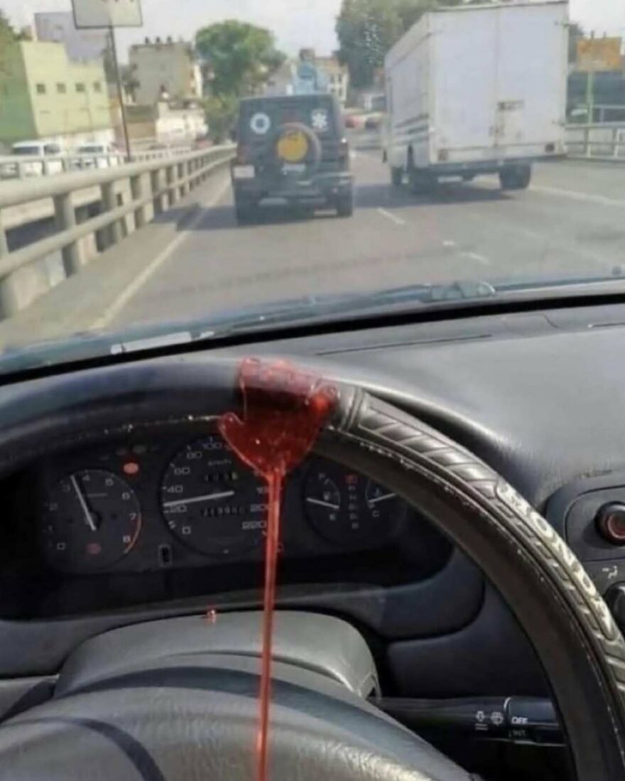 Steering wheel covered in a red liquid resembling blood inside a moving car, bizarre and hilarious roadside view.