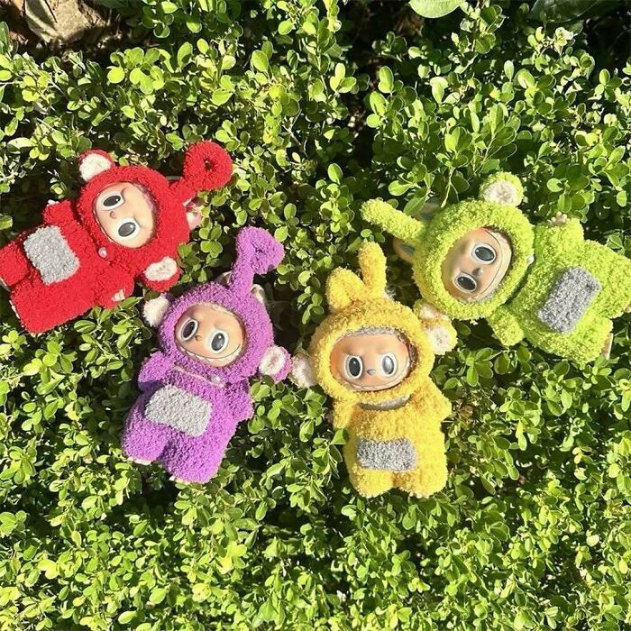 Colorful plush Teletubby toys in red, purple, yellow, and green laid on green leafy background, cool secondhand finds.