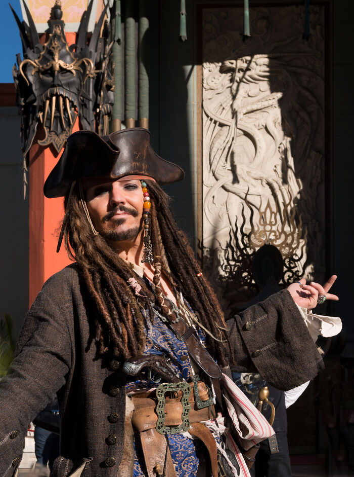 Man dressed as pirate with dreadlocks and leather hat, posing outdoors near intricate historic architectural details.