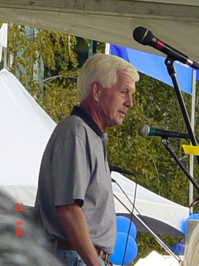 Older man speaking at an outdoor event with microphones nearby, symbolizing late bloomers becoming millionaires.