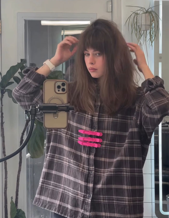Young woman with depression hairstyle taking a mirror selfie, capturing the trendy Gen Z hair look in casual plaid shirt.
