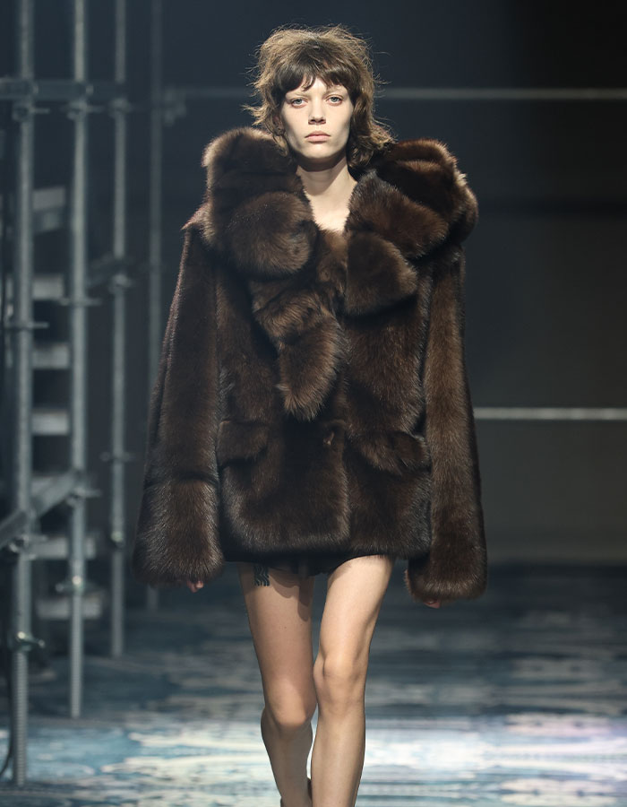 Model with depression hairstyle walking runway wearing oversized brown fur coat during fashion show.