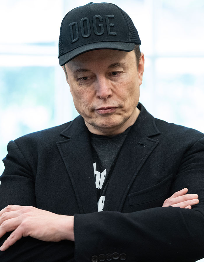 Elon Musk wearing a black DOGE cap and black blazer, looking down with a serious expression showing signs of aging.