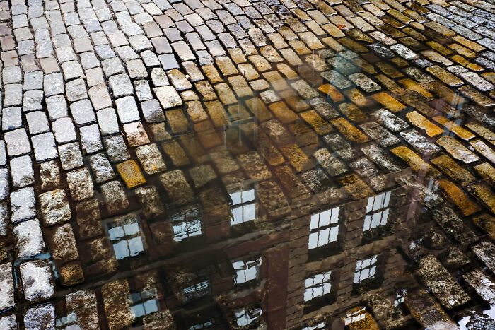 Reflection of a city building on wet cobblestone street, showcasing street photographer’s magical city life coincidences.