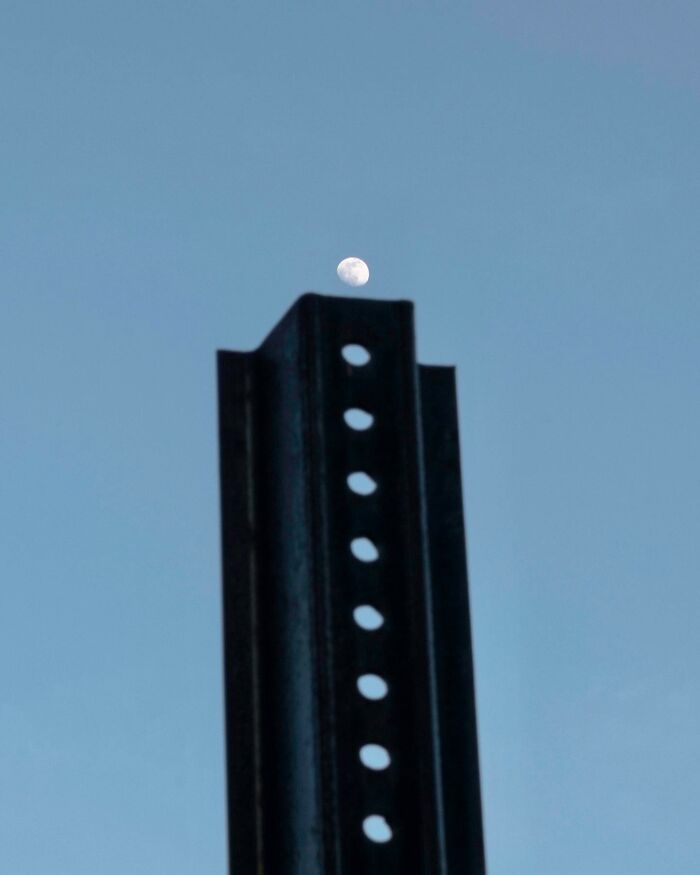 Street photographer captures a coincidence where the moon perfectly aligns with a metal post against a clear sky.