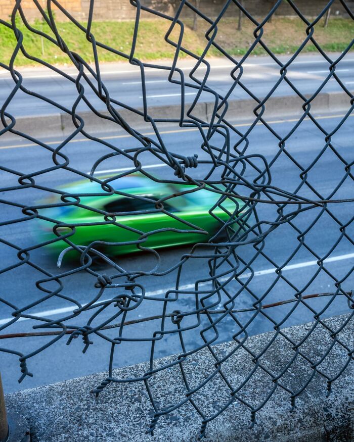 Green car blurred in motion seen through a broken wire fence, capturing a street photographer’s magical city life coincidence.
