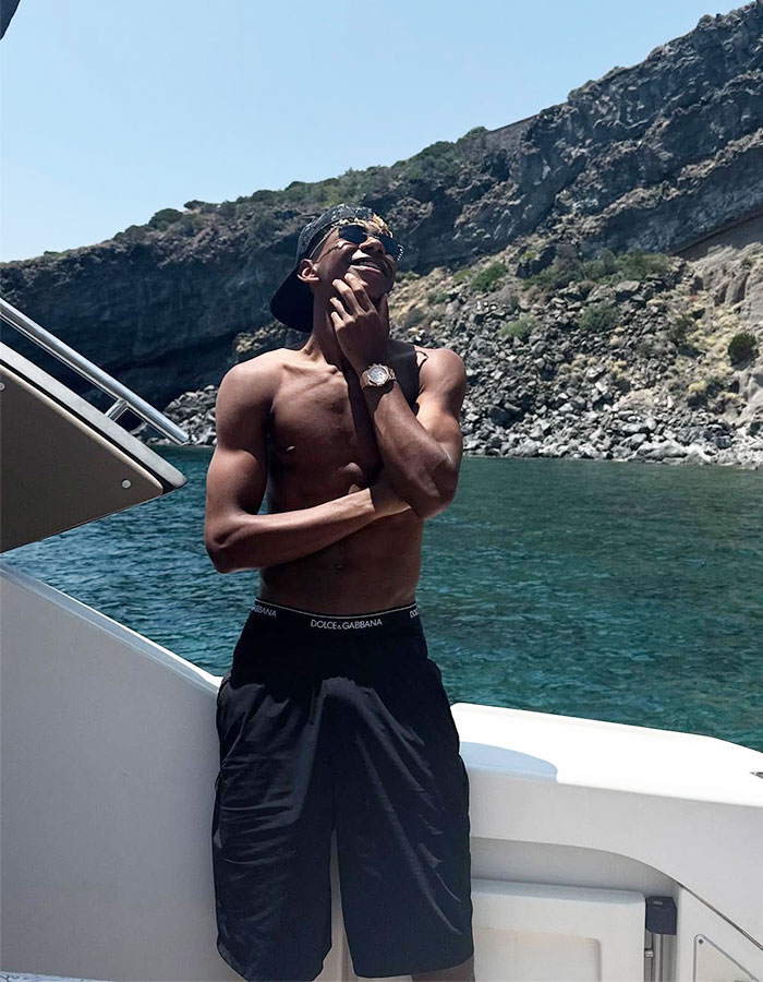 Influencer 29 enjoying vacation on boat near rocky coastline, wearing sunglasses and shorts, soaking in the sun.