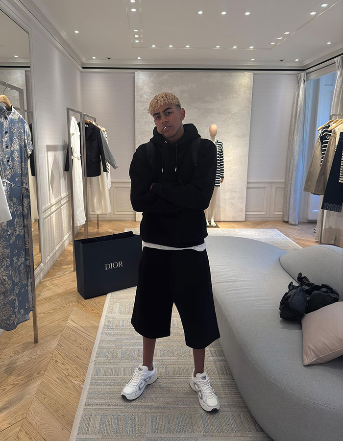 Influencer standing in a luxury boutique wearing black hoodie and shorts, amid clothing racks and a Dior shopping bag.