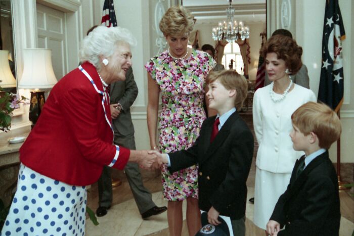 Princess Diana in a floral dress watching two boys shake hands with an older woman during a formal royal event indoors.