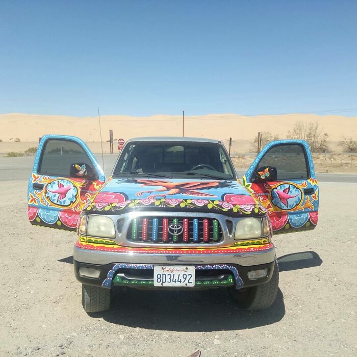 Toyota truck painted with colorful, intricate patterns featuring birds and flowers in a desert setting with open doors.