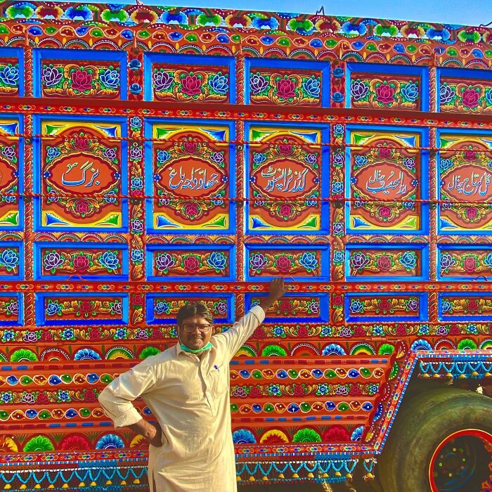 Man posing in front of a truck decorated with colorful, intricate patterns as works of art on the vehicle's surface.