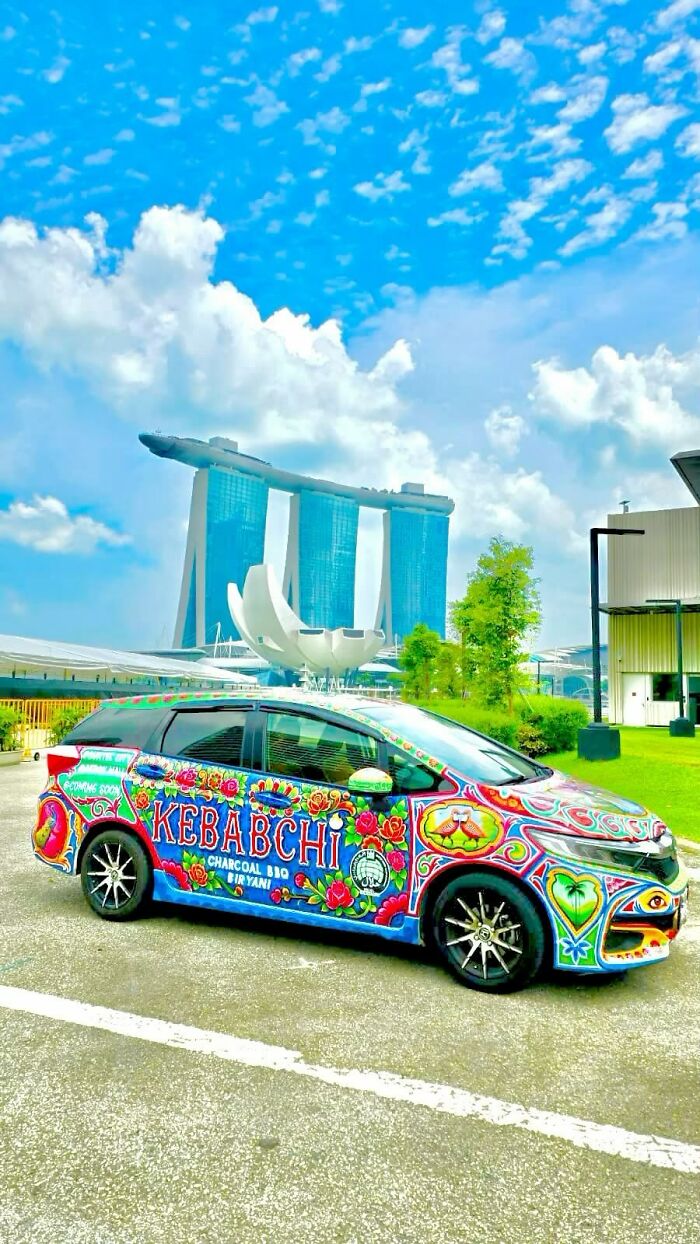 A car covered in colorful, intricate patterns and vibrant artwork parked outdoors with a bright blue sky background.