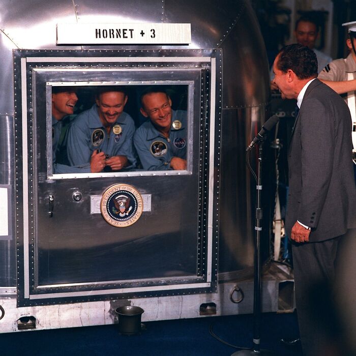 Apollo mission astronauts smiling inside quarantine cabin while a man in suit speaks into microphone nearby