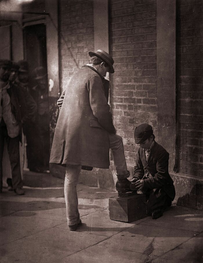 Victorian times street scene showing a boy shining shoes for a man in a long coat and hat on a city sidewalk.