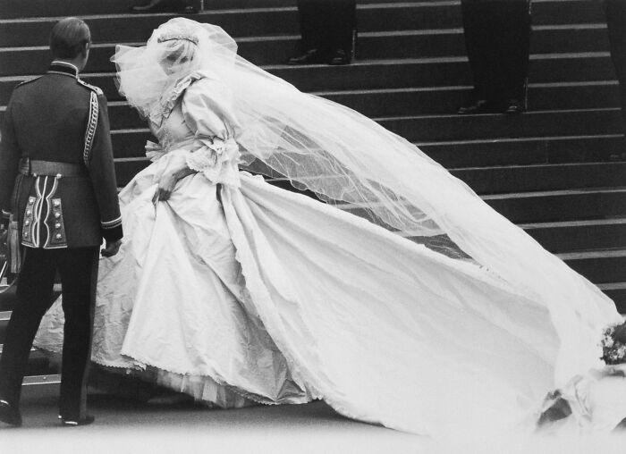 Princess Diana wearing a flowing wedding dress and veil ascending steps beside a man in formal military attire.