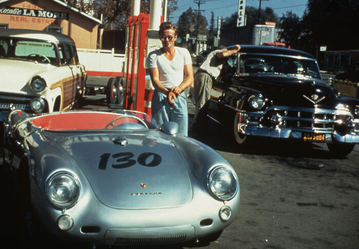 Young man standing by a vintage Porsche at a gas station, one of the last known photos of famous people.