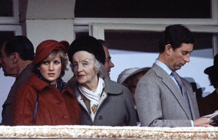 Princess Diana wearing a red coat and hat standing beside an elderly woman and a man in a gray suit reading a program.