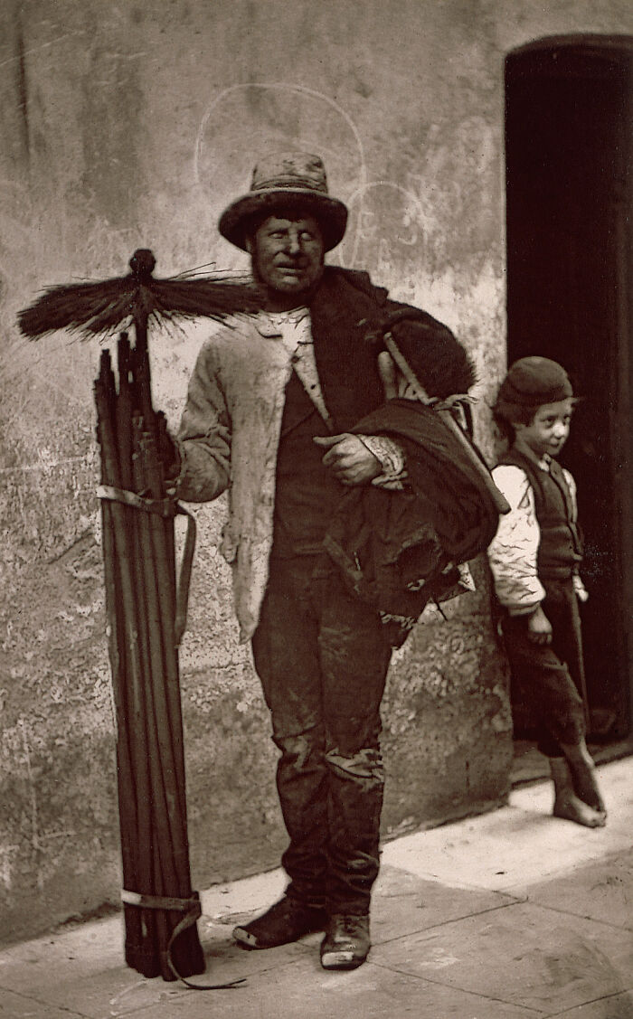 Victorian times street scene showing a man holding cleaning tools and a young boy standing near a doorway.