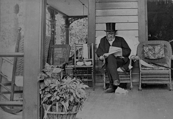 An elderly man wearing a top hat sitting on a porch reading a newspaper, one of the last known photos of famous people.