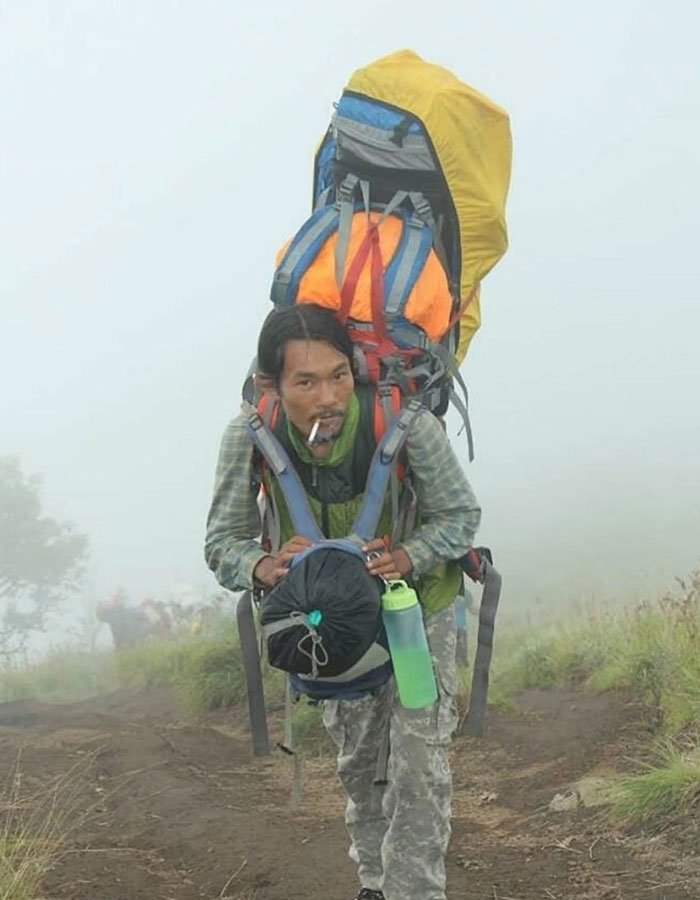Mountain guide carrying large backpack climbing foggy trail during active volcano body recovery mission.