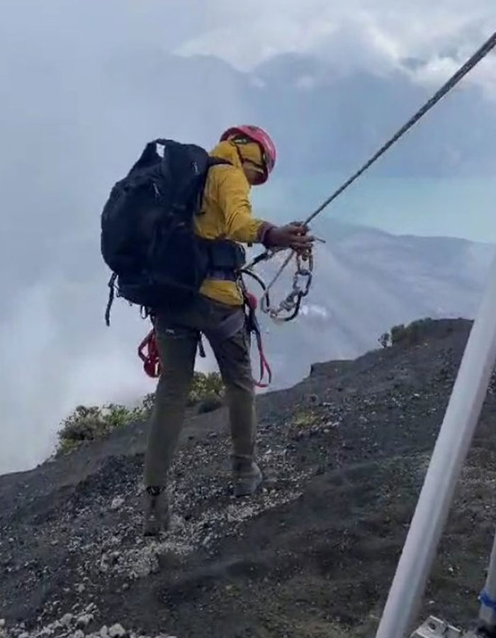 Rescuer in yellow jacket and helmet descending active volcano with rope during body recovery from 1600ft-deep site.