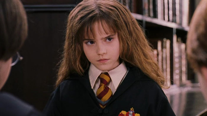 Young Hermione Granger in Hogwarts uniform looking serious during a Harry Potter scene in the magical library setting.