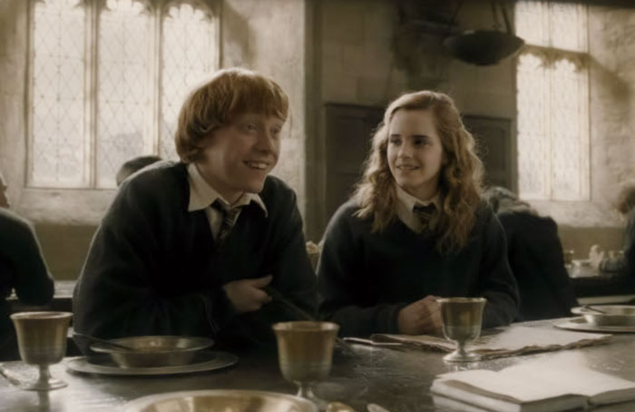Emma Watson and co-star sitting at a Hogwarts dining table in a Harry Potter film scene.