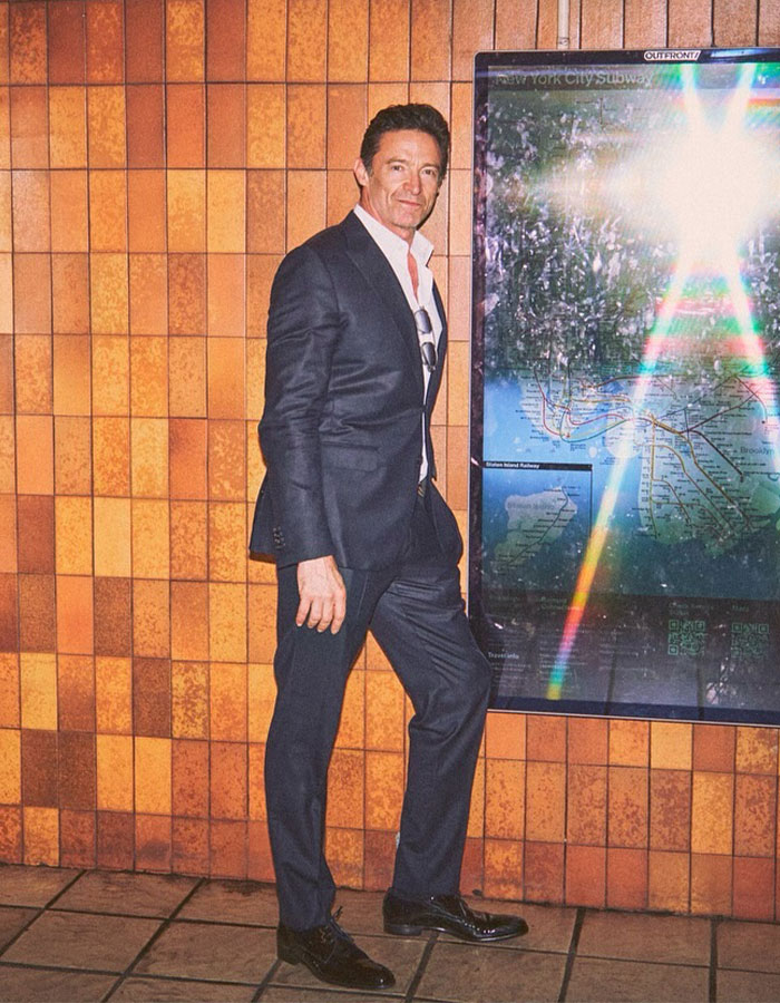 Hugh Jackman in a dark suit standing near a subway map, reflecting light, amid divorce setback rumors.