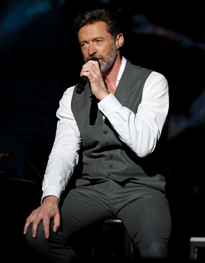 Hugh Jackman holding microphone during performance, wearing grey vest and white shirt, amid divorce setback news.