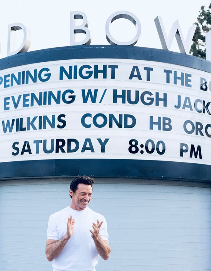 Hugh Jackman standing and clapping in front of a theater marquee featuring his name in bold letters.