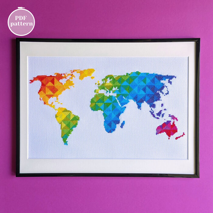 Framed colorful cross-stitched map of the world with geometric patterns on white fabric against a purple background.