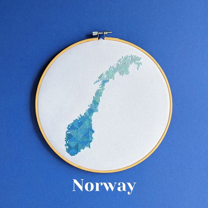 Cross-stitched map of Norway in shades of blue and green displayed in an embroidery hoop on a blue background.