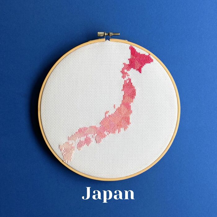 Cross-stitched map of Japan in shades of pink and red displayed in an embroidery hoop on a blue background.
