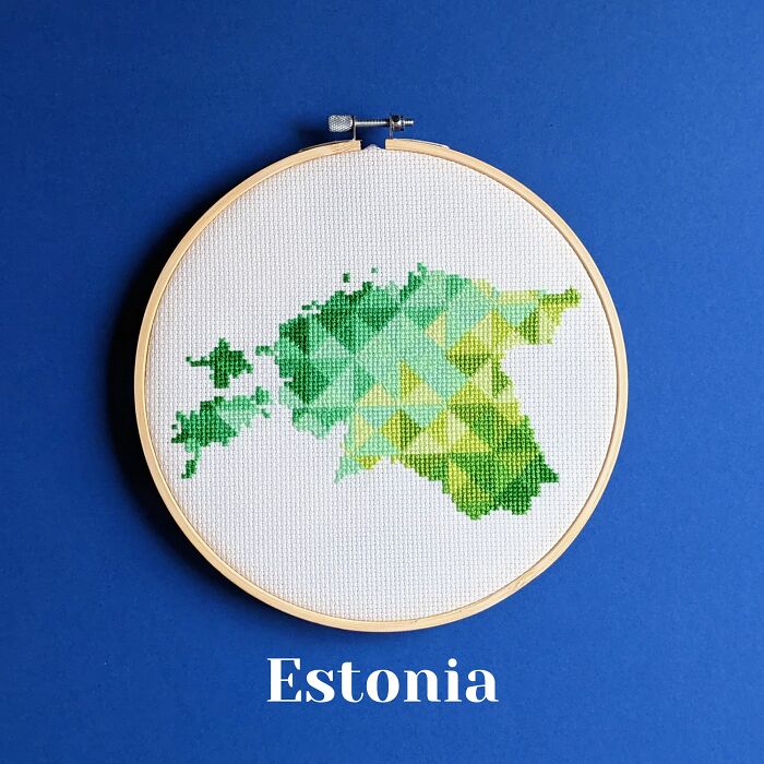 Cross-stitched map of Estonia in green shades displayed in a wooden embroidery hoop on a blue background.