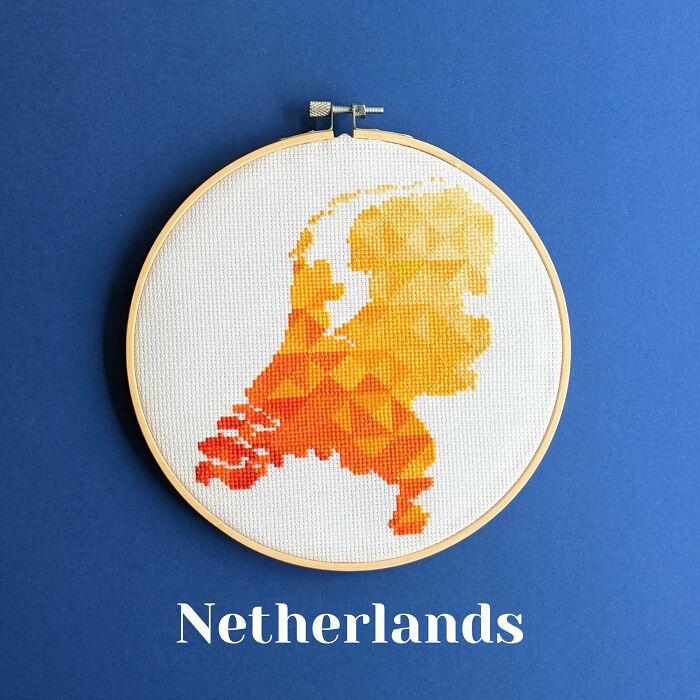 Cross-stitched map of the Netherlands in warm orange tones displayed in an embroidery hoop on a blue background.