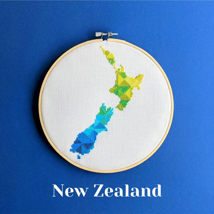 Cross-stitched map of New Zealand with colorful geometric patterns in an embroidery hoop on a blue background.