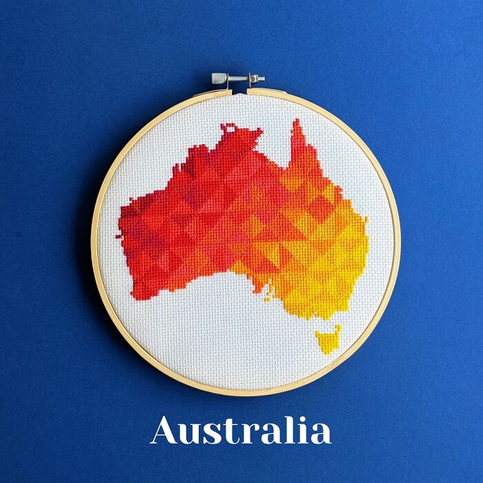 Cross-stitched map of Australia in warm red and yellow tones displayed in an embroidery hoop on a blue background.