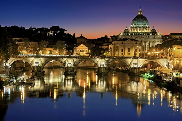 Night view of Rome with illuminated bridge and dome reflecting on the river, showcasing world capitals geography knowledge.